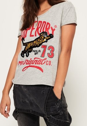 Nainen, joka pukeutunut harmaaseen t-paitaan, jossa on hyppivän tiikerin grafiikka ja punainen teksti "Superdry 73 Original Co." yhdistettynä mustiin kulutettuihin farkkushortseihin.