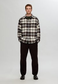 Checkered flannel shirt in beige and dark brown with two chest pockets, paired with dark brown trousers and black shoes.