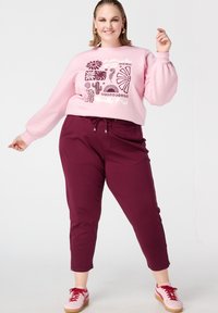 Lichtroze sweatshirt met grafisch patroon, gecombineerd met bordeauxrode joggerbroek met verstelbare taille. Afgewerkt met roze sneakers.