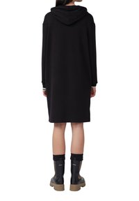 Schwarzes Kapuzenkleid in lockerer Passform, mit langen Ärmeln und kontrastierenden Bündchen. Das Modell trägt schwarze Stiefel mit einem hellbraunen Sohlen.