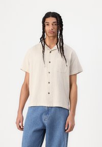 Short-sleeve cream button-up shirt with a textured fabric, single chest pocket, and gray buttons, paired with loose-fitting blue jeans.