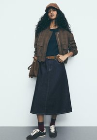 Ženska s kodrasto lasmi, oblečena v rjav karirast suknjič in kapo, temnobluzo, denim midi krilo, rjav pas, drži rjav nahrbtnik, nosi športne čevlje in maroon nogavice.