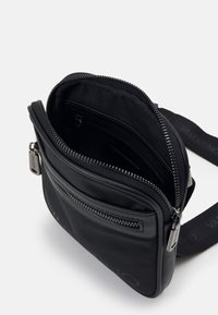 Černá crossbody taška se zipovými kapsami, vyrobená z nylonu a kůže. Má nastavitelný popruh a detail s logem na přední straně.