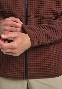 Braune gesteppte Jacke mit hohem Kragen, vollem Reißverschluss und enganliegenden Bündchen. Der Stoff hat eine erhabene Struktur mit einem Rautenmuster. Minimalistisches Design.