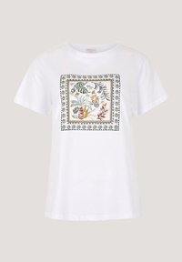 T-shirt bianca in cotone con stampa floreale quadrata nei toni del verde, giallo e marrone, bordata da un motivo decorativo nero.