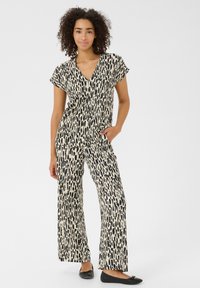 Combinaison à motifs noirs et crème avec des manches courtes, un décolleté en V, une coupe ample et un pantalon large. Le mannequin pose les mains dans les poches.