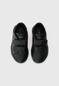 Mustat nahkakengät, joissa on kaksi Velcro-nauhaa, kumipohja, pyöreä kärki ja ommellut yksityiskohdat. Sisällä on "Lacoste" -merkintä.