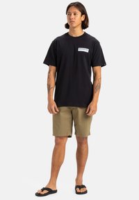 T-shirt en coton noir avec un petit logo blanc sur la poitrine, associé à un short kaki et des tongs noires. Aucun motif ou texture notable.