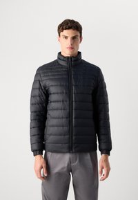 Young man wearing a black quilted jacket with a front zipper and gray pants, standing against a plain white background.
