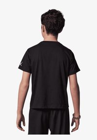 Czarny t-shirt z bawełny, krótki rękaw, okrągły dekolt i subtelny detal z logo na lewym ramieniu. Widok z tyłu pokazuje luźny krój.