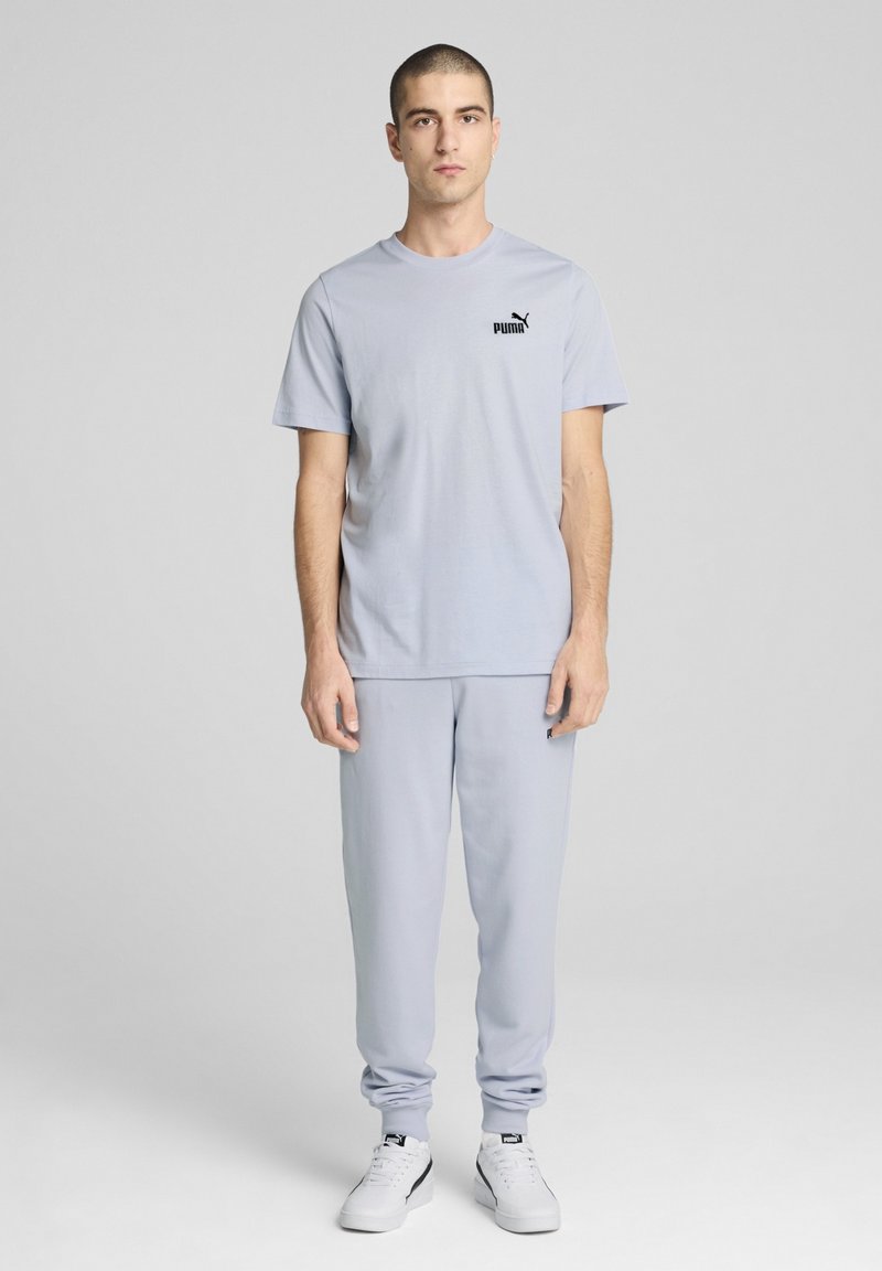 Set de îmbrăcăminte sportivă asortată în gri deschis, compus dintr-un tricou cu mânecă scurtă și pantaloni jogger, confecționat dintr-un material moale, având un accent cu logo negru pe piept.