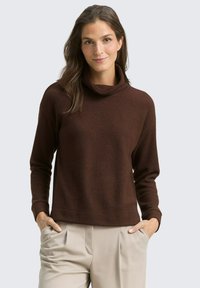 Pull en maille marron à col cheminée, avec des manches longues et une coupe décontractée. Porté avec un pantalon beige, les mains dans les poches. Fond uni.