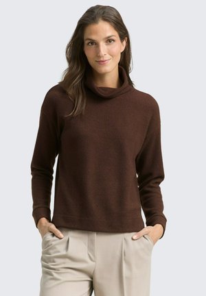 Pull en maille marron à col cheminée, avec des manches longues et une coupe décontractée. Porté avec un pantalon beige, les mains dans les poches. Fond uni.