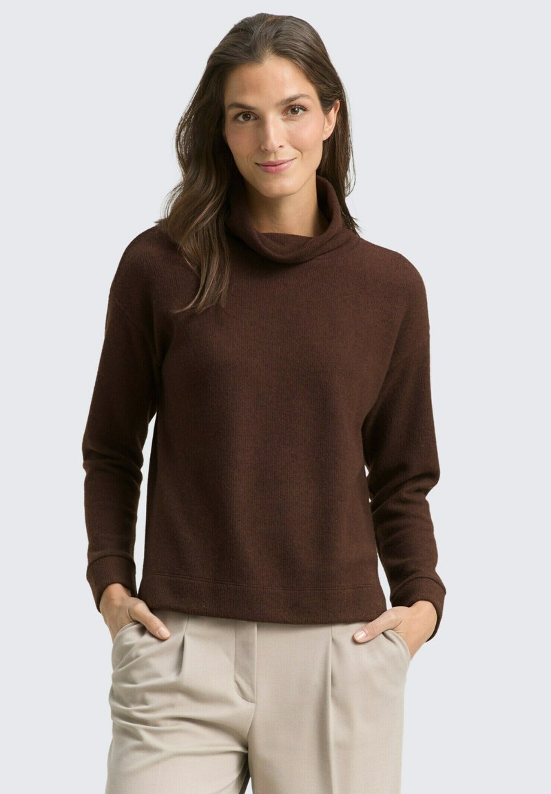 Pull en maille marron à col cheminée, avec des manches longues et une coupe décontractée. Porté avec un pantalon beige, les mains dans les poches. Fond uni.