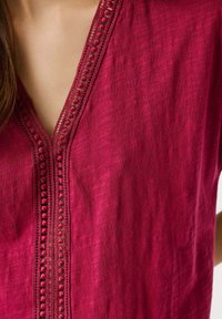 Close-up of a red garment with decorative crochet trim along a V-shaped neckline and part of a woman's bare shoulder visible.