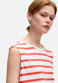 Weißes Shirt mit roten horizontalen Streifen, kurzen Ärmeln, rundem Ausschnitt und weichem Material. Model mit kurzen Haaren und Ohrringen in Form von Ringen.