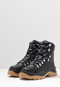 Botas de senderismo de cuero negro con suelas de goma marrón resistentes, ojales de metal plateado y cordones en negro y blanco con un patrón entrecruzado.