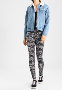 Giubbotto in denim con colletto in shearling bianco, abbinato a leggings a fantasia bianca e nera, e sneaker nere. Vista a figura intera.