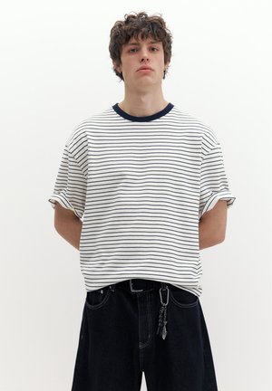 Ung mand med krøllet hår iført en hvid kortærmet skjorte med sorte horisontale striber og sorte bukser med nøglering som tilbehør.