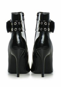 Zapatos de tacón alto de charol negro con tacón de aguja, que presentan una amplia correa para el tobillo, ojales plateados y una textura suave y brillante.