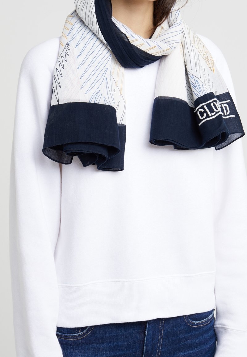 Femme portant un sweat-shirt blanc et un jean bleu avec une écharpe blanche et marine présentant des motifs de lignes abstraites et un logo à une extrémité.