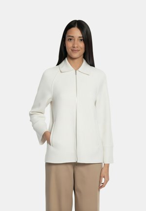 Donna con lunghi capelli scuri che indossa una giacca bianca con cerniera e collo e pantaloni beige, in piedi con una mano in tasca.