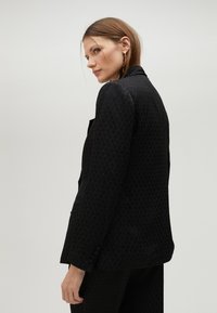 Černý blazer se strukturovaným vzorem, moderním střihem a dlouhými rukávy. Obsahuje klopový límec a boční kapsy, s knoflíkovými detaily na rukávech.