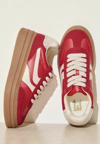 Raudoni ir balti retro sportbačiai, turintys zomšos ir odos viršutinę dalį, apvalias raištelius, storą gumos pado sluoksnį ir tekstūruotą šoninio rašto dizainą.