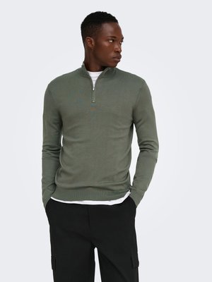 Homme portant un pull vert olive à fermeture éclair quart sur une chemise blanche et un pantalon noir, les mains dans les poches, regardant sur le côté sur un fond uni.