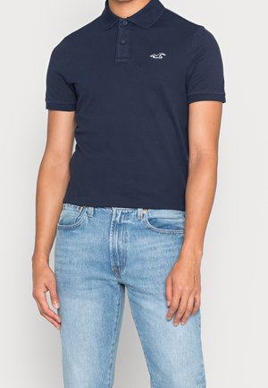 Homme portant un polo bleu marine à manches courtes avec un petit logo blanc et un jean bleu clair, debout les bras détendus.