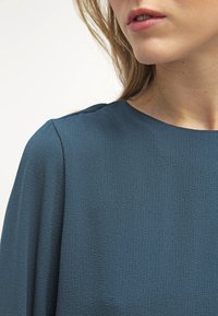 Turkusowa bluzka z miękkiego, teksturowanego materiału, z okrągłym dekoltem i subtelnym drapowaniem na ramionach, prezentująca gładkie wykończenie.