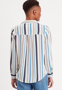 Blouse à manches longues en tissu léger avec des rayures verticales bleu marine, bleu et crème. Présente un col et des poignets à boutons.