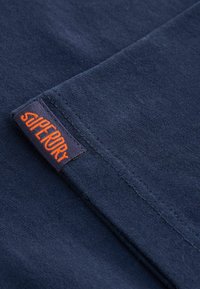 Mörkblå bomullstyg med en mjuk yta, som har en synlig orange "Superdry"-etikett på sömmen. Tyget är välsytt.
