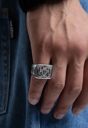 Inel din metal argintiu cu un cadru pătrat, decorat cu modele intricate de leu și coroană. Mâna prezintă o textură a pielii în contrast cu materialul din denim.