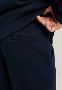 Granatowe dresy o miękkiej fakturze, z tylną kieszenią i ribbowanym elastycznym paskiem w talii. Ręka ciągnąca za pas, aby odsłonić materiał.