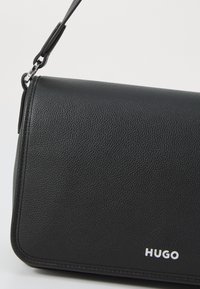 Svart skinncrossbody-veske med en teksturert overflate, rektangulær form, sølvfarget hardware og logodetalj nederst til høyre.