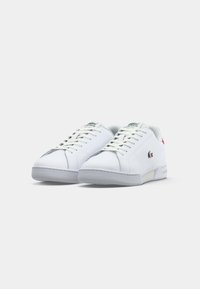 Par bijelih Lacoste niskih tenisica s vezicama, brandski logo na strani i jeziku, te suptilne sive soles protiv jednostavne bijele pozadine.