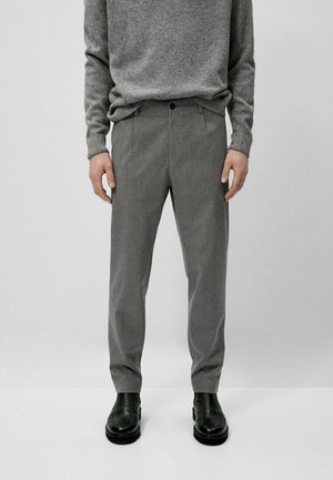 Man wearing grey wool sweater and grey tailored trousers with black leather boots, standing against a plain light background.