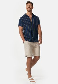 Lachende man die loopt, gekleed in een blauw gestreept overhemd met korte mouwen, beige korte broek en witte sandalen met gesp.