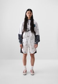 White oversized hoodie with black detailing and signature prints, paired with a gray and black apron-style bottom and red-accented shoes.
