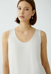 Haut sans manches blanc avec un décolleté en rond, texture lisse et détails de couture minimaux. Le mannequin porte des boucles d'oreilles en perles.