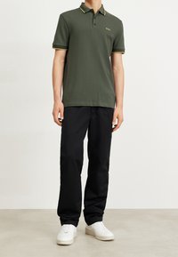 Mörkgrön pikétröja med gula accenter på kragen och ärmarna, bärs med svarta byxor och vita sneakers. Enkelt och avslappnat design.