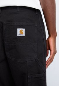 Black canvas pants featuring a large back pocket with a white label displaying a yellow logo and brand name, with reinforced stitching details.