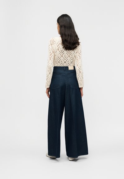 Γυναίκα με μακριά σκουρόχρωμα μαλλιά στέκεται με την πλάτη, φοράει μπεζ πουλόβερ με ανοιχτή πλέξη και σκούρα τζιν παντελόνια με φαρδιά πόδια, μπροστά από ένα απλό φόντο.