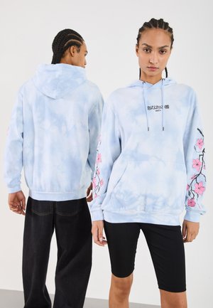 Hoodie legat, de culoare bleu deschis, cu un model tie-dye, dotat cu un buzunar frontal, broderie florală roz pe mâneci și cu un glugă cu șnur. Este asortat cu pantaloni închiși la culoare și pantaloni scurți negri.