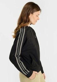 Kvinna i svart zip-up jacka med vita ränder på ärmarna, händerna i fickorna, vänd mot höger mot en enkel ljus bakgrund.