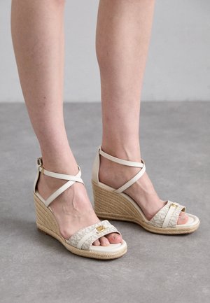 Sandalias de cuña beige con tiras de tela texturizada, diseño de cruz en el tobillo y tacón de yute trenzado. Presenta detalles en dorado en la tira.