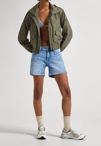 Olivgrön kort jacka, ljusblå denimshorts, grå sneakers med vita detaljer, beige strumpor; enkel design och avslappnad stil.