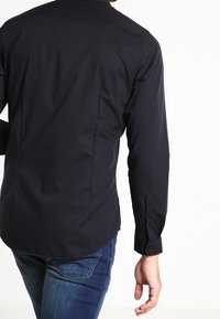 Black long-sleeve shirt with a button-down collar, featuring a smooth texture and fitted design, paired with blue denim jeans.