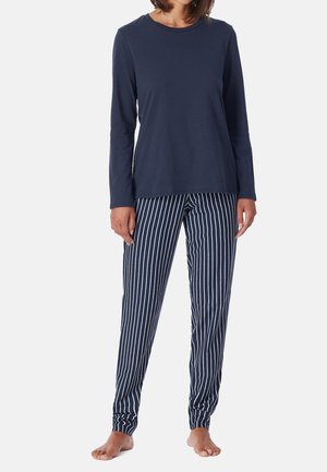 Kobieta ubrana w granatową bluzkę z długim rękawem i granatowe spodnie w paski, stojąca boso na jasnym, jednolitym tle.